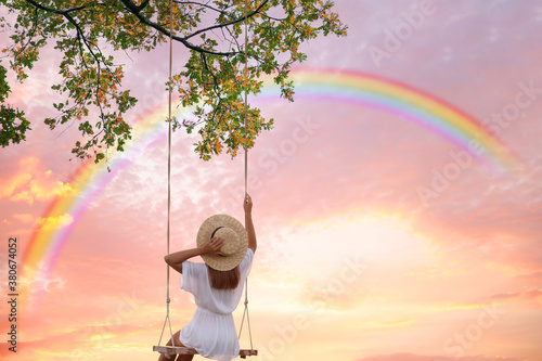 Dream world. Young woman swinging, rainbow in sunset sky on background photo