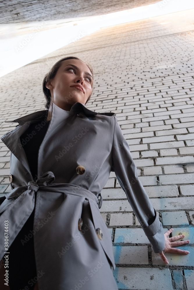 gorgeous asian attractive model wearing trench coat standing near wall shot from lower point. outdoors photoshoot. portrait of young beautiful sensual female
