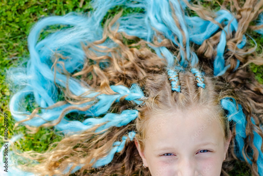 Portrait of beautiful girl with many braids with blue kanekalon.Cute girl 7 years old with braids on green grass background. girl laying on a lawn. Festive, modern hairstyle