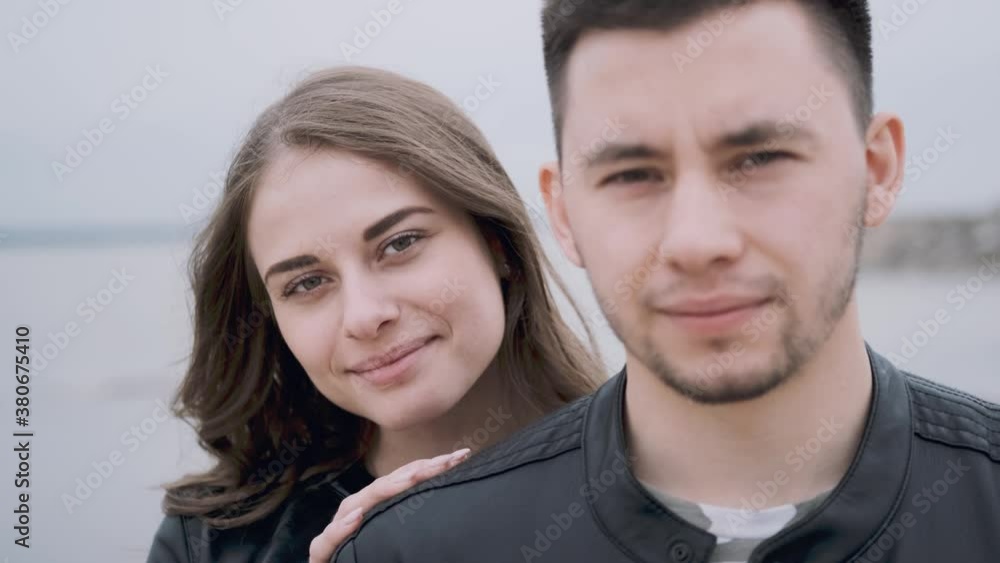 Close-up portrait of a happy young couple at the seaside on a cloudy day. A beautiful girl hugs her boyfriend from the back. The camera zooms out slowly. Friendship and love concept. Close-up portrait of a happy young couple at the seaside on a cloudy day. A beautiful girl hugs her boyfriend from the back. The camera zooms out slowly. Friendship and love concept.