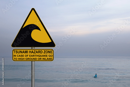 Tsunami warning and evacuation sign located on a beach. Blue calm sea misty landscape and only one boy floating on the surf board at smooth water as background 