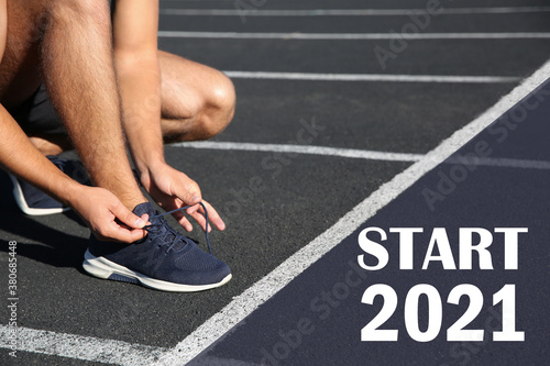 Sporty man tying shoelaces in front of starting line with text Start 2010 at stadium, closeup