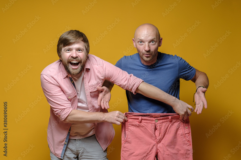 Obraz premium Two friends are shopping. Cheerful man jokingly tried on large pants for another perplexed men. Sale concept. Yellow background.
