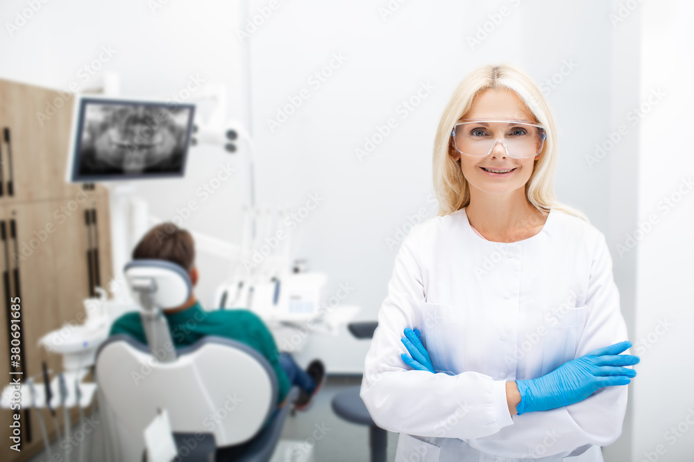 Portrait of a woman dentist smiling with crossed hands, background dentistry clinic with a patient. Teeth treatment modern clinic Portrait of a woman dentist smiling with crossed hands, background dentistry clinic with a patient. Teeth treatment modern clinic