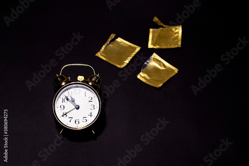 A golden watch is on a black background. There're three opened condoms behind the watch. Special design for the contraception day. There is a space for text.