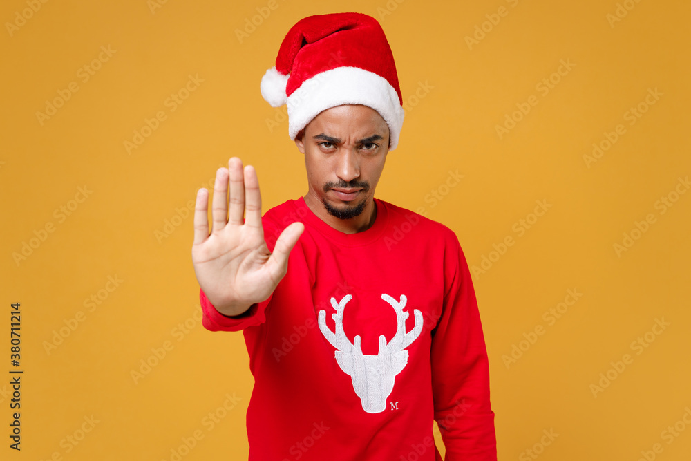 Dissatisfied young Santa african american man 20s in red sweater Christmas hat showing stop gesture with palm isolated on yellow background studio portrait. Happy New Year celebration holiday concept.