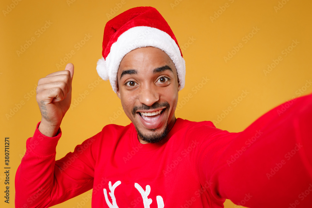 Close up of happy young Santa african american man in Christmas hat doing selfie shot on mobile phone doing winner gesture isolated on yellow background. Happy New Year celebration holiday concept. Close up of happy young Santa african american man in Christmas hat doing selfie shot on mobile phone doing winner gesture isolated on yellow background. Happy New Year celebration holiday concept.