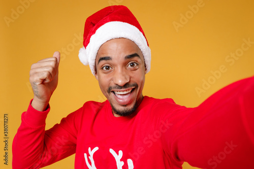 Close up of happy young Santa african american man in Christmas hat doing selfie shot on mobile phone doing winner gesture isolated on yellow background. Happy New Year celebration holiday concept. © ViDi Studio