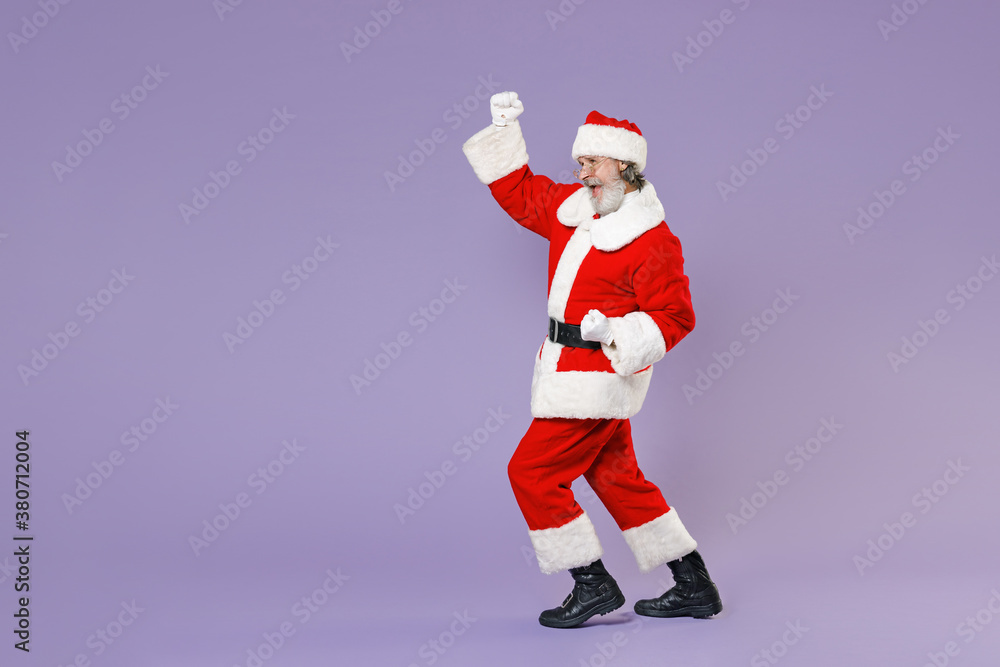 Full length side view portrait Santa Claus man in Christmas hat red suit coat white gloves glasses doing winner gesture isolated on violet background. Happy New Year celebration merry holiday concept. Full length side view portrait Santa Claus man in Christmas hat red suit coat white gloves glasses doing winner gesture isolated on violet background. Happy New Year celebration merry holiday concept.