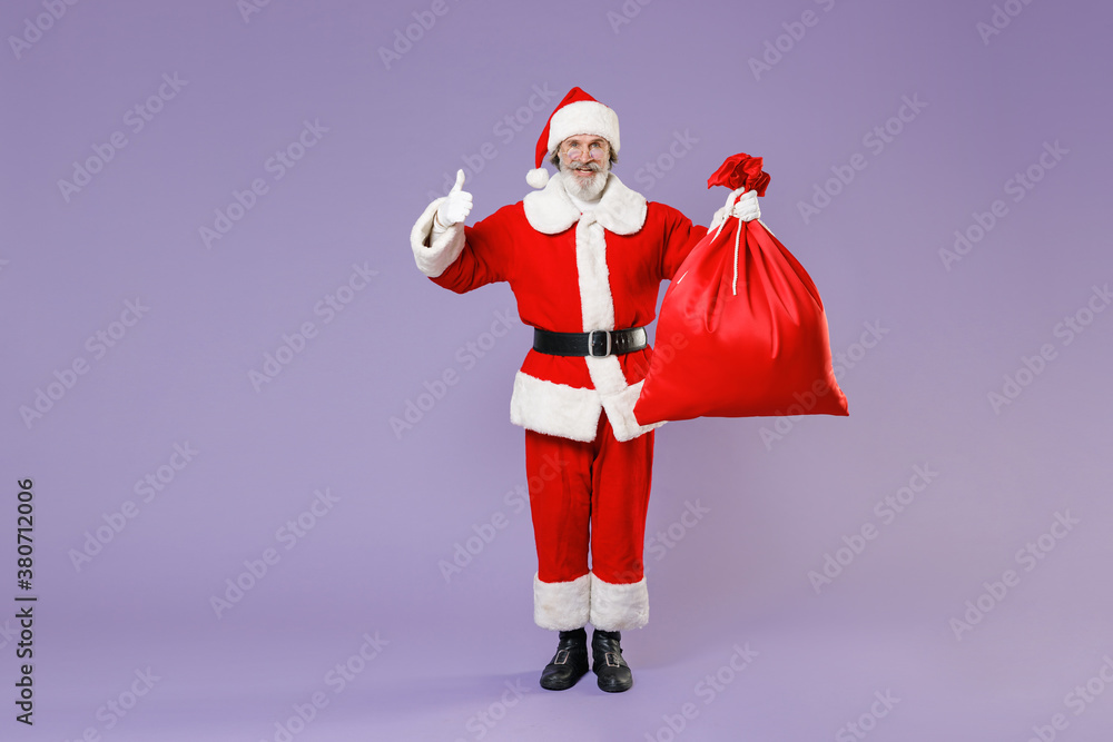 Full length portrait of funny Santa Claus man in Christmas hat coat suit hold bag with presents gifts showing thumb up isolated on violet background. Happy New Year celebration merry holiday concept. Full length portrait of funny Santa Claus man in Christmas hat coat suit hold bag with presents gifts showing thumb up isolated on violet background. Happy New Year celebration merry holiday concept.