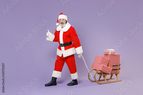 Full length portrait excited Santa Claus man in Christmas hat suit carries sleigh with present gifts boxes showing thumb up isolated on violet background. Happy New Year celebration holiday concept.