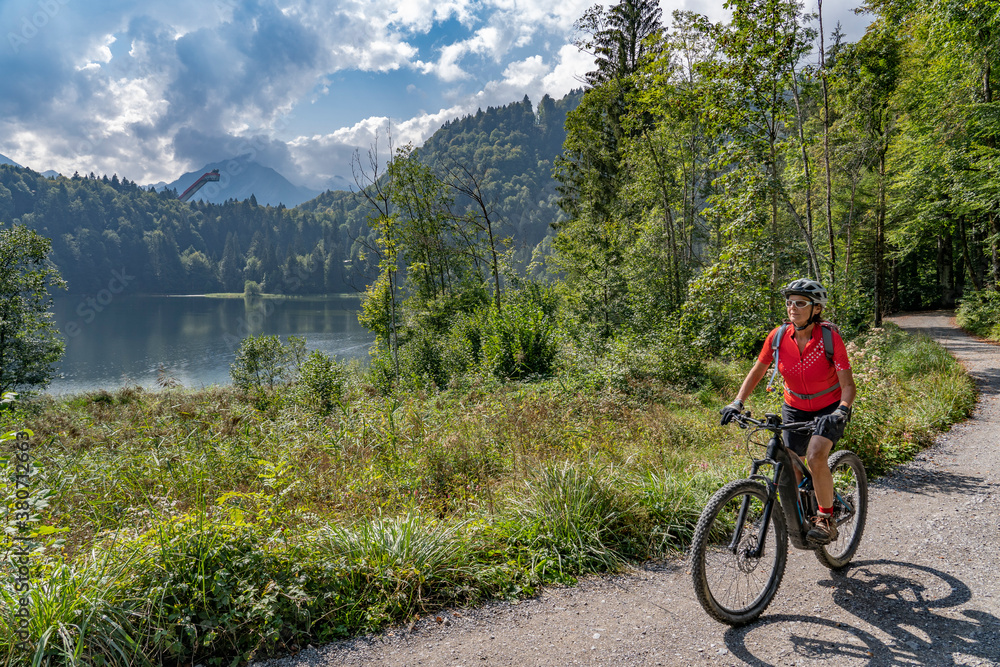 Fototapeta premium nice senior woman riding her electric mountain bike above the Freiberg Lake and a big ski flying hill in the Allgau Alps near Oberstdorf, Bavaria, Germany 