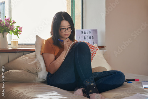 Girl in glasses drawing in notebook on bed.