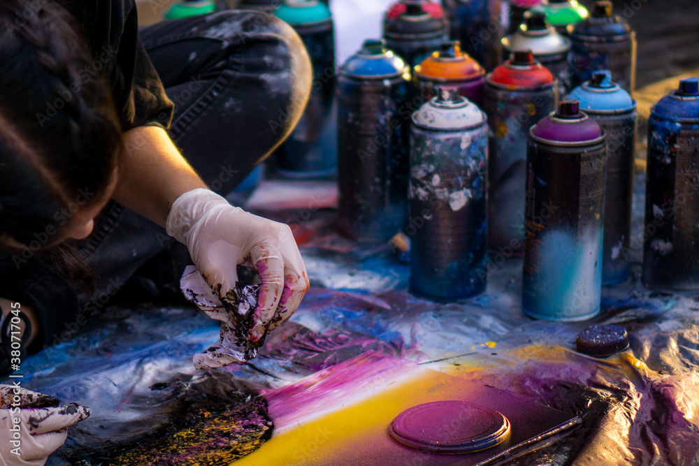 Fototapeta premium girl street artist in gloves paints with spray cans of paint on a piece of paper. multicolored cans of paint stand around. copy space