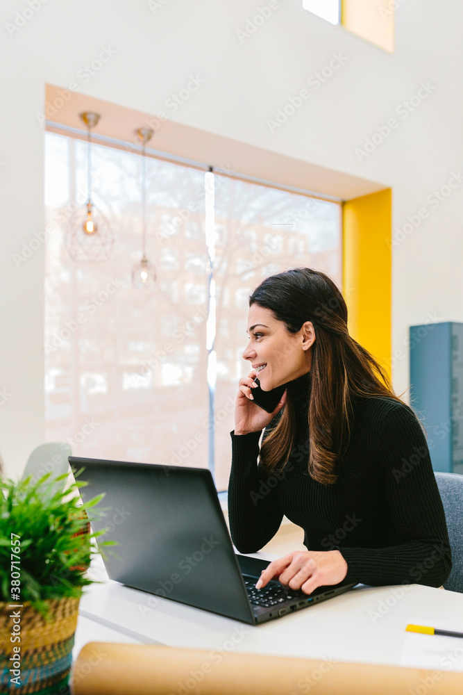 Smiling woman having phone call sitting at desk with laptop Smiling woman having phone call sitting at desk with laptop