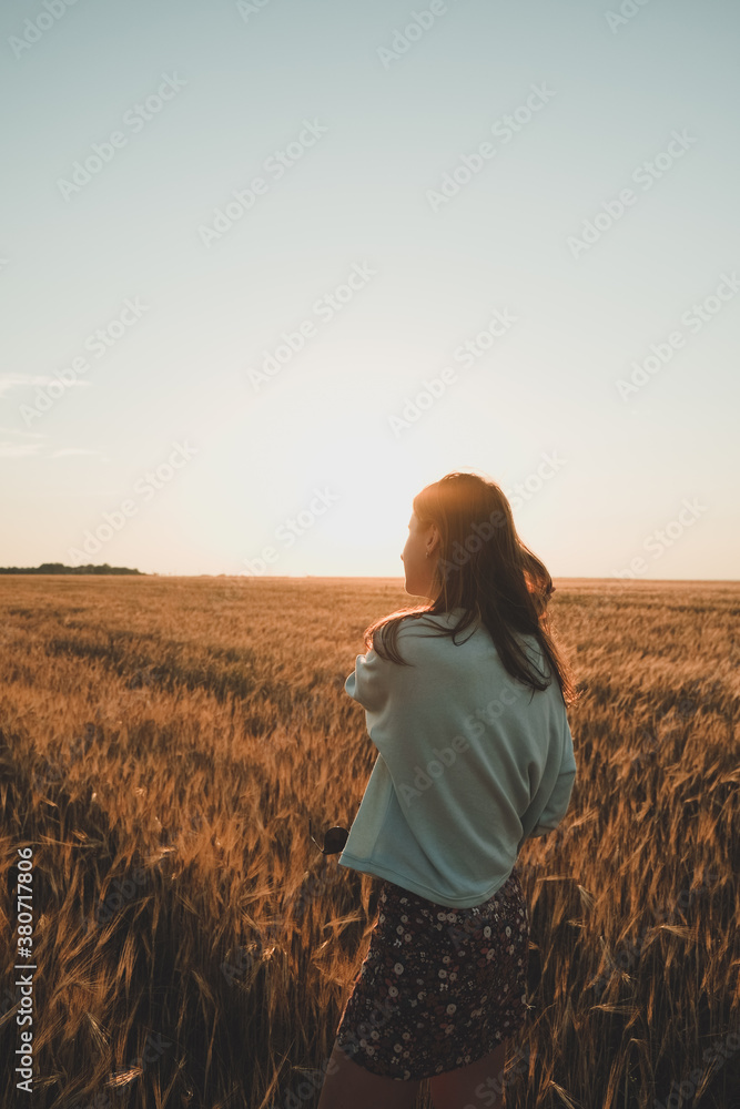 Fototapeta premium Amazing Girl on the wheat field at sunset. Nature landscape with dramatic clouds sunset sky. Summer background. Rear view, contour light. Travel concept