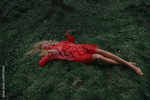 top view of a beautiful long-legged model with blond hair lying on a green moss in the forest and looking at the sky. on her red translucent raincoat
