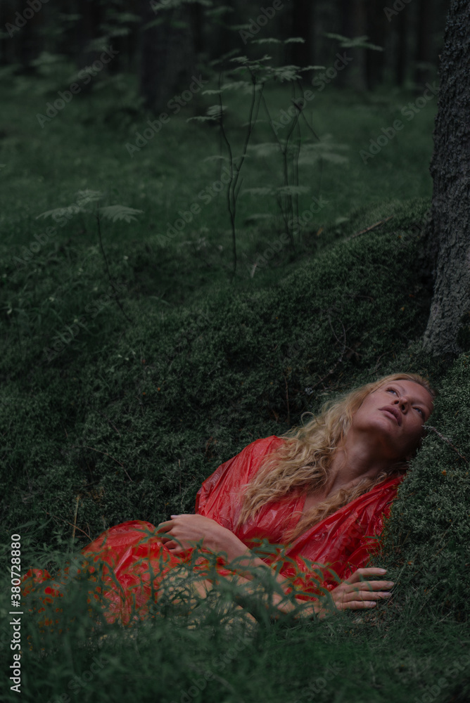 beautiful blonde woman in a red translucent raincoat lies in a moss under a tree and looks up beautiful blonde woman in a red translucent raincoat lies in a moss under a tree and looks up
