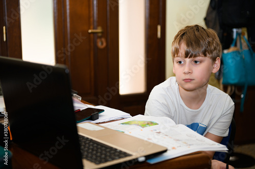 The boy looks longingly at the computer screen. Homeschooling during the coronavirus. E-education, cyberbullying concept, tired or sad student boy with laptop computer sitting near a desk at home 