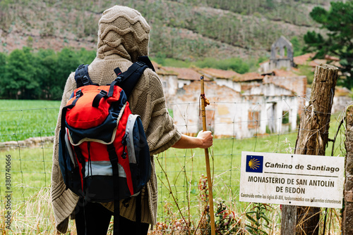 Back view of unrecognizable hiker with backpack and trekking stick standing against ancient monastery of San Antolin de Bedon during pilgrimage on Camino de Santiago in Spain photo