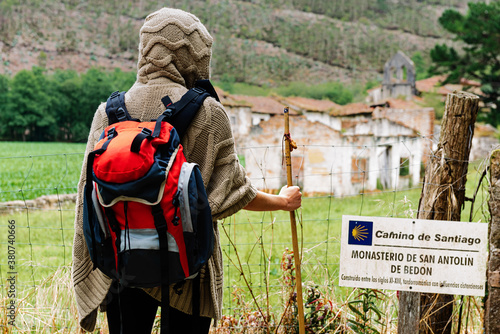 Back view of unrecognizable hiker with backpack and trekking stick standing against ancient monastery of San Antolin de Bedon during pilgrimage on Camino de Santiago in Spain