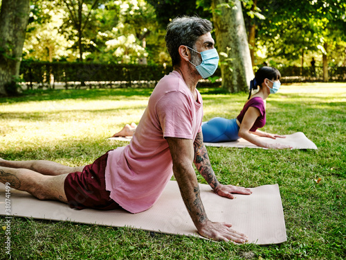 Side view of serene couple in protective masks practicing yoga in Bhujangasana on mats in green park in summer during coronavirus outbreak photo