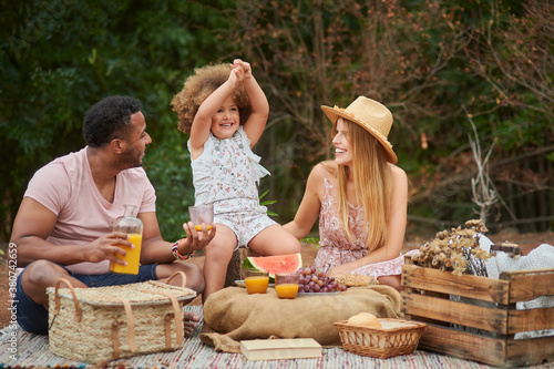 Loving ethnic dad pouring natural orange juice into glass during picnic with wife and little daughter in summer day in nature photo