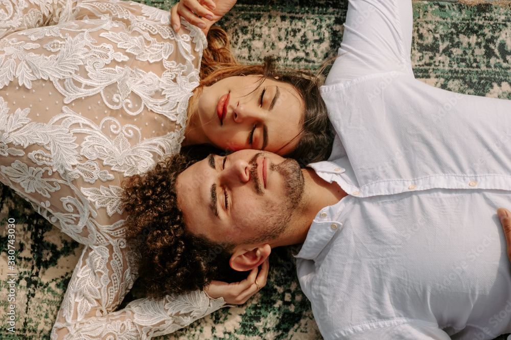 From above romantic newlywed young couple in stylish clothes lying opposite with face to face with eyes closed cuddling on vintage rug on sandy beach From above romantic newlywed young couple in stylish clothes lying opposite with face to face with eyes closed cuddling on vintage rug on sandy beach