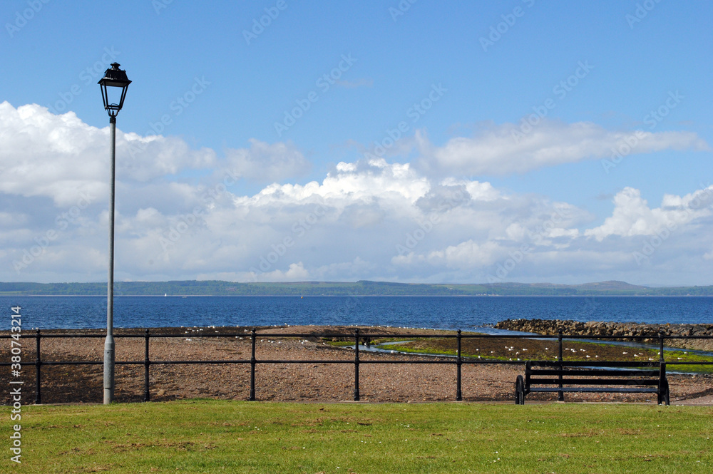 Isolated Metal Lamppost & Empty Bench on Grassy Promenade beside Seashore against Blue Sky