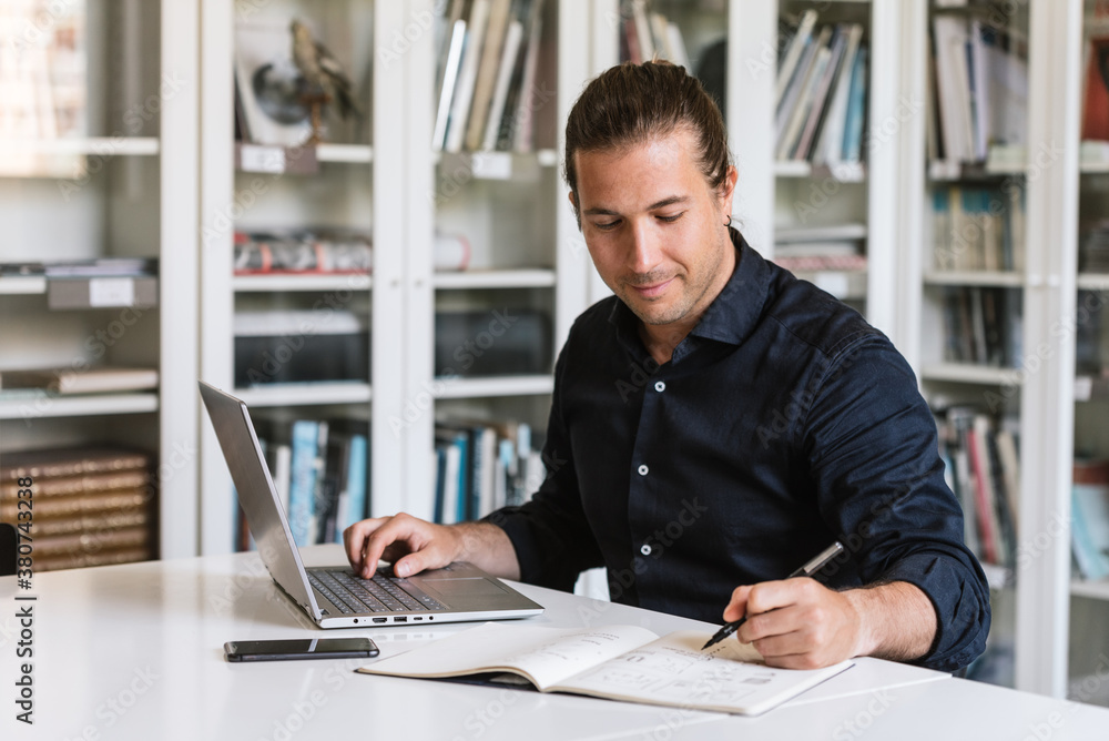 Focused businessman in formal shirt sitting at table while doing paperwork and typing on notebook in bright workspace Focused businessman in formal shirt sitting at table while doing paperwork and typing on notebook in bright workspace