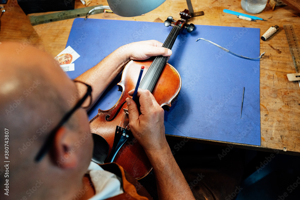 High angle back view of artisan craftsman adjusting strings on violin bridge while working at wooden desk in workshop High angle back view of artisan craftsman adjusting strings on violin bridge while working at wooden desk in workshop
