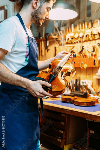 Side view of young craftsman in apron standing with shiny violin near workbench in modern workshop photo