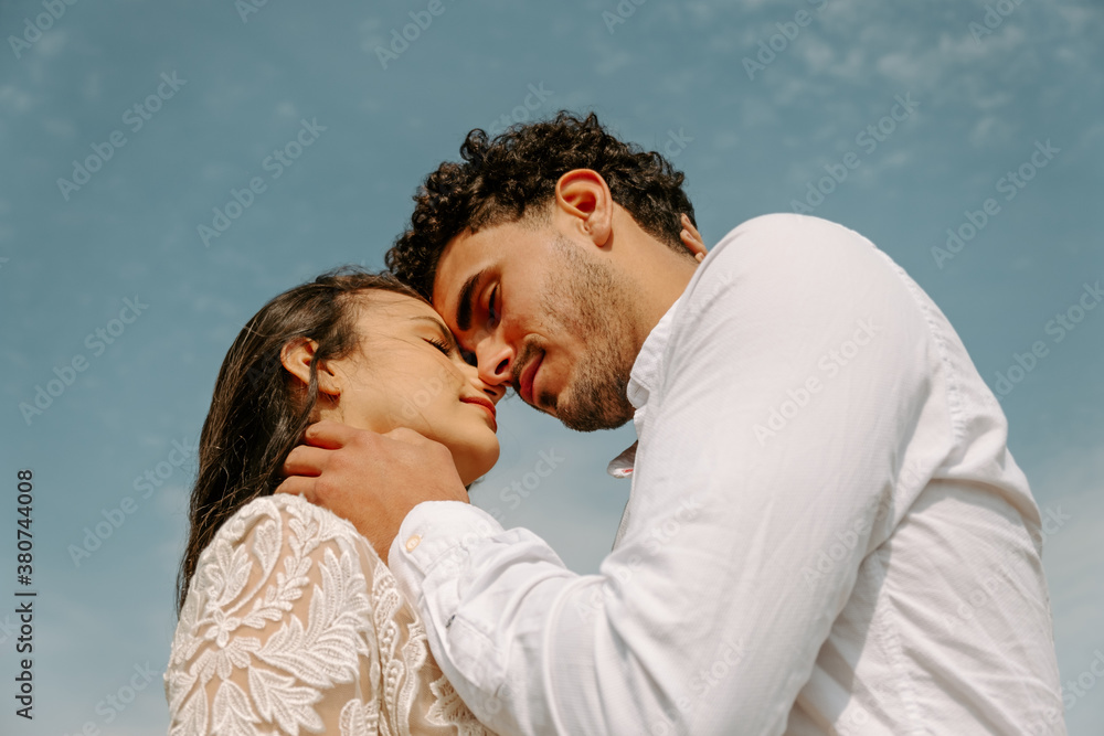 Low angle side view of happy young newlywed couple in white wedding outfits hugging and kissing against blue sky Low angle side view of happy young newlywed couple in white wedding outfits hugging and kissing against blue sky