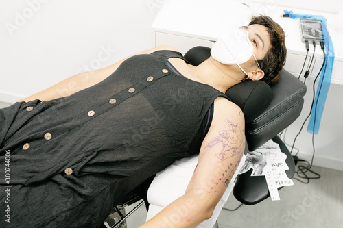 Calm female in dress and protective mask lying on table and preparing for getting tattoo in salon