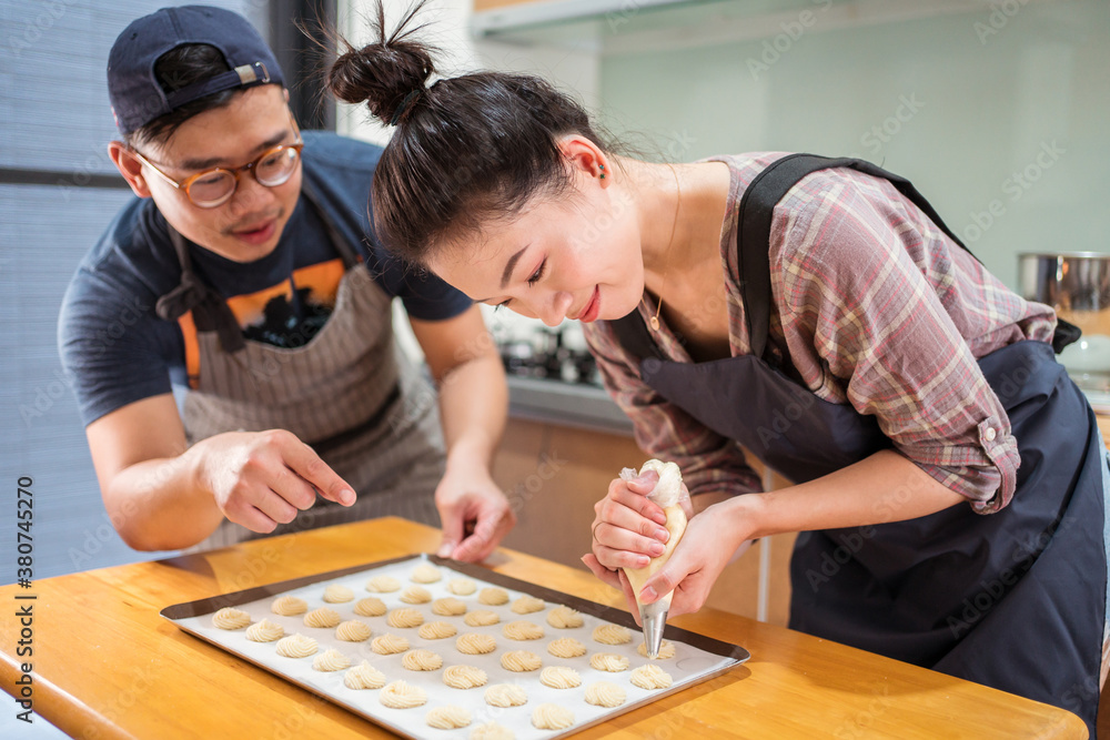 Asian guy helping smiling girlfriend with piping bag to pipe batter for pastries on baking pan at counter Asian guy helping smiling girlfriend with piping bag to pipe batter for pastries on baking pan at counter