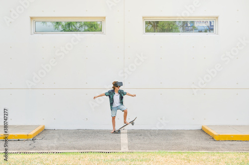 Kid in summer clothes doing a trick with a skateboard wearing virtual reality glasses on a parking in the street photo