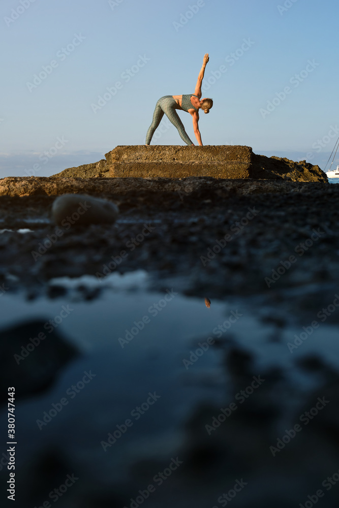 Side view of slim female in sportswear doing Revolved Triangle asana while practicing yoga on rocky seashore Side view of slim female in sportswear doing Revolved Triangle asana while practicing yoga on rocky seashore