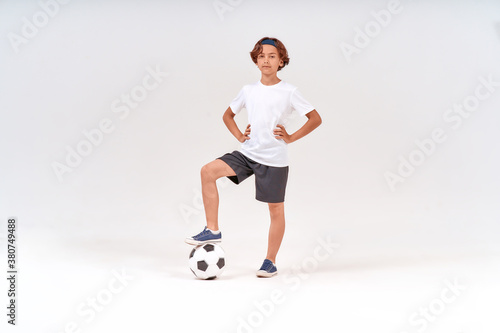 Football player. Full-length shot of a happy teenage boy with soccer ball looking at camera and smiling while standing isolated over grey background © Kostiantyn