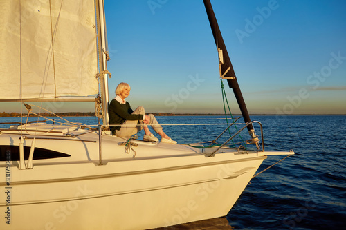 Enjoying luxury life. Beautiful senior woman sitting on the side of sailboat or yacht deck floating in the calm blue sea at sunset, looking away and smiling