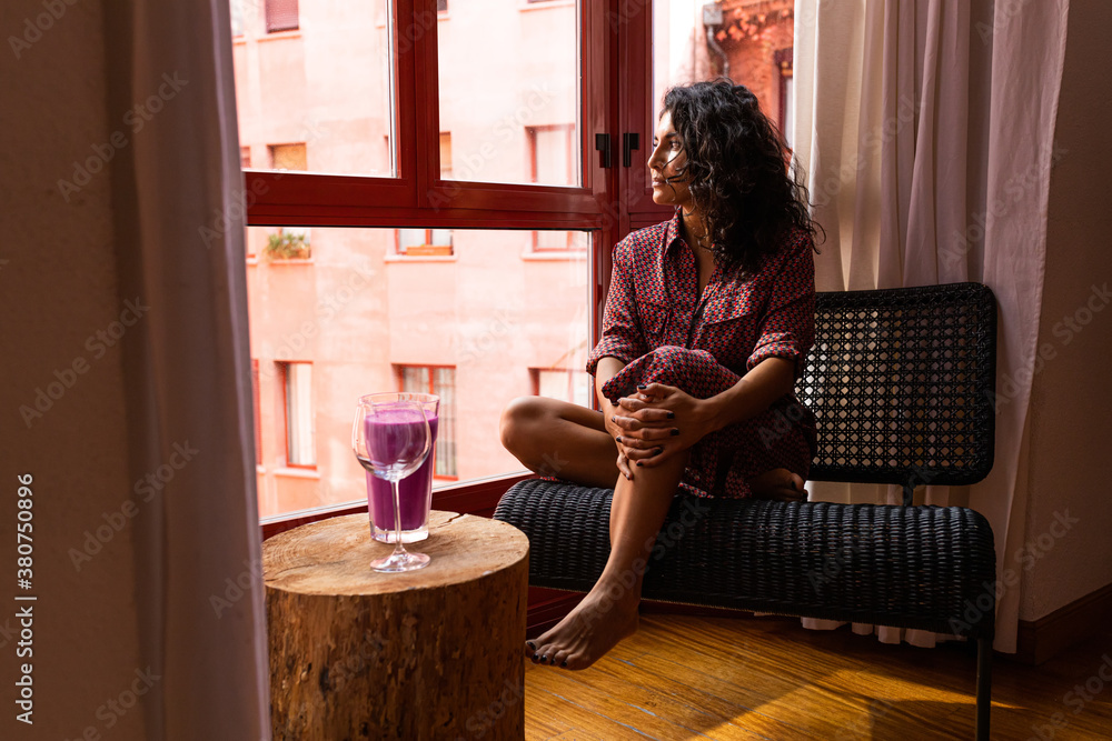 Ethnic thoughtful latin woman enjoying a healthy kefir and pitaya smoothie sitting by the windows at home Ethnic thoughtful latin woman enjoying a healthy kefir and pitaya smoothie sitting by the windows at home