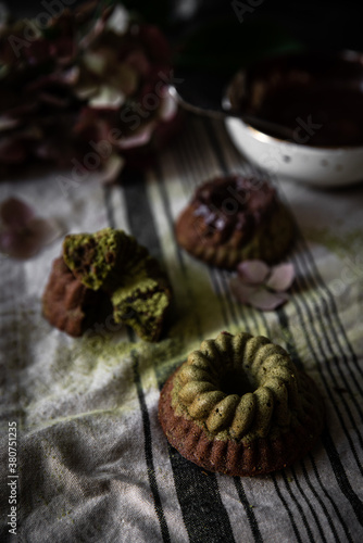 Mini chocolate-matcha bundt cakes, bowl with chocolate and hydrangea flowers on linen towel on dark background.