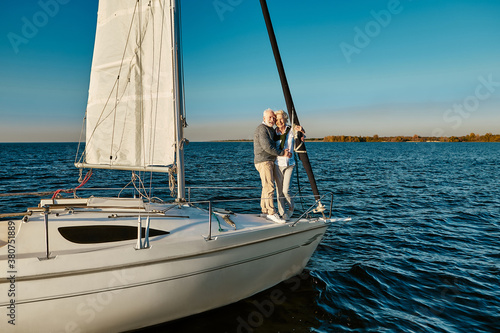 Boat trip. Happy senior man and woman hugging and enjoying amazing view while standing on the side of sail boat or yacht deck floating in the calm blue sea, sailing together