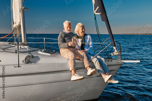 Celebrating wedding anniversary. Happy senior couple drinking wine or champagne and laughing while sitting on a sailboat or yacht deck floating in a calm blue sea