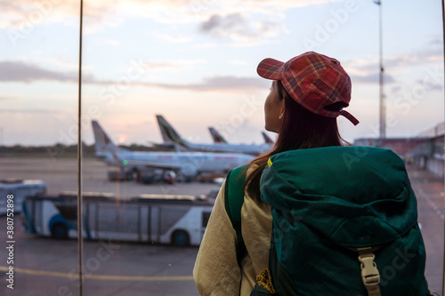 Back view of young Asian female traveler with backpack standing near window in airport terminal and observing airplanes while waiting for flight in Sri Lanka