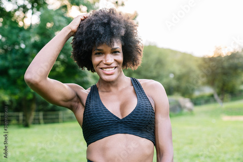 Young African American sportswoman with curly hair wearing black top looking at camera with smile while standing in green park during workout photo