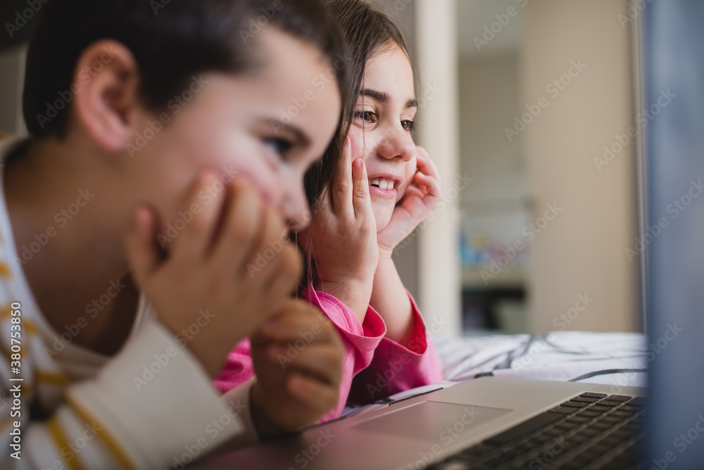 Happy little sister and brother having fun and playing game on laptop together while spending free time in bedroom at home Happy little sister and brother having fun and playing game on laptop together while spending free time in bedroom at home