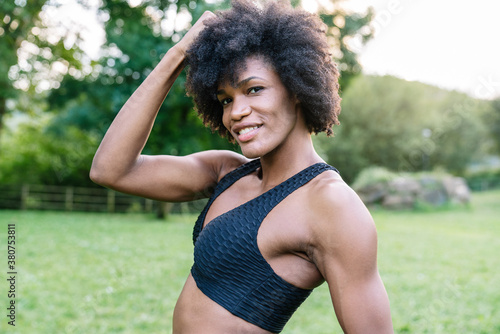 Young African American sportswoman with curly hair wearing black top looking at camera with smile while standing in green park during workout photo