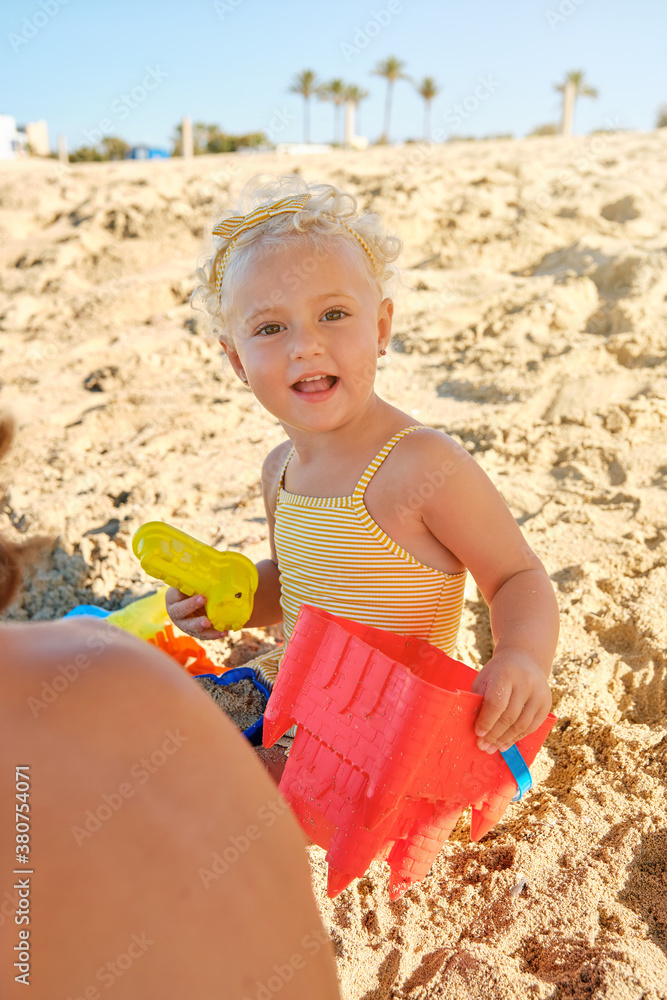 Vertical photo of a little blonde girl with a yellow striped swimsuit sitting on the sand playing to fill plastic toys with sand under the shadow while facing the camera on the beach