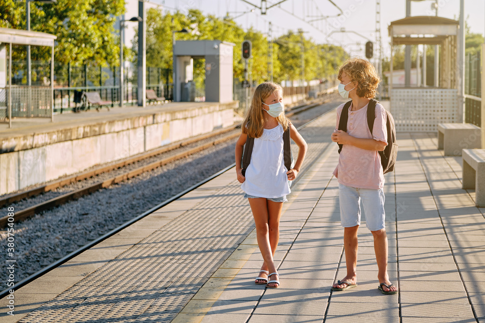 Two children with black school bags and mask waiting for the train on a platform at the station in the morning Two children with black school bags and mask waiting for the train on a platform at the station in the morning