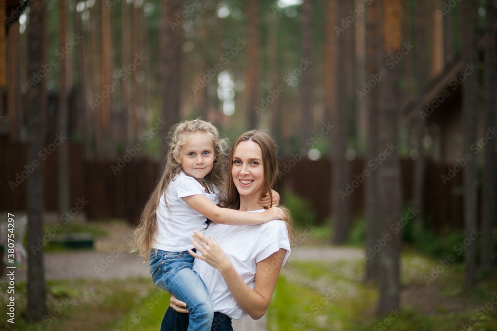 Fototapeta premium Happy smiling mother and her cute emotional little daughter child in white shirts and denim jeans are hugging and having fun outdoor in nature at countryside. Spending summer time with family.